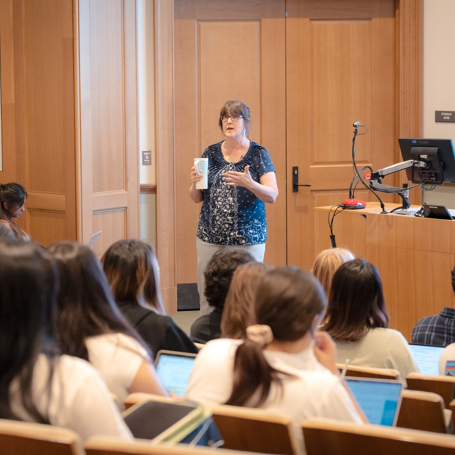 A UP professor holds white mug while standing near a wooden wall lecturing a class. Students are partially visible in the lower left corner. The setting appears to be indoors, with warm wood paneling.