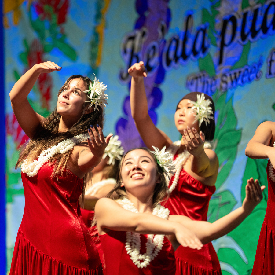 A University of Portland Hawaii Club member with flowers in her hair raises one arm and looks upward, performing a traditional dance in front of a colorful, abstract background.