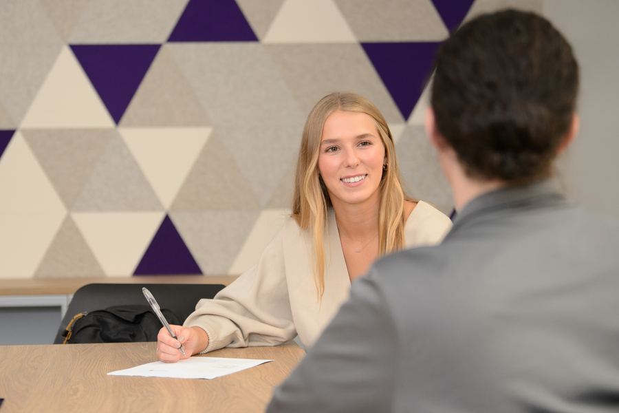 An image showing the back of a persons head with dark hair speaking to a UP student with blond hair set against a background featuring a purple triangle shape.