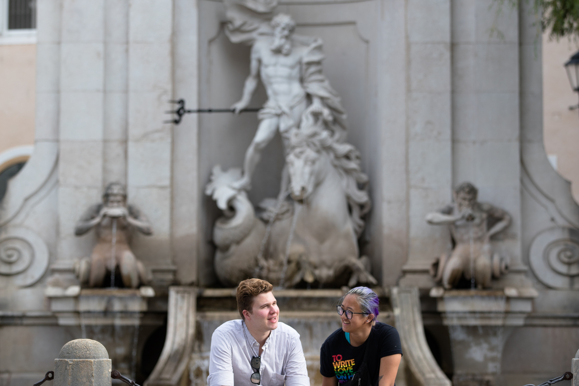Two UP students are chatting casually while seated in front of a classical sculpture in a European plaza.