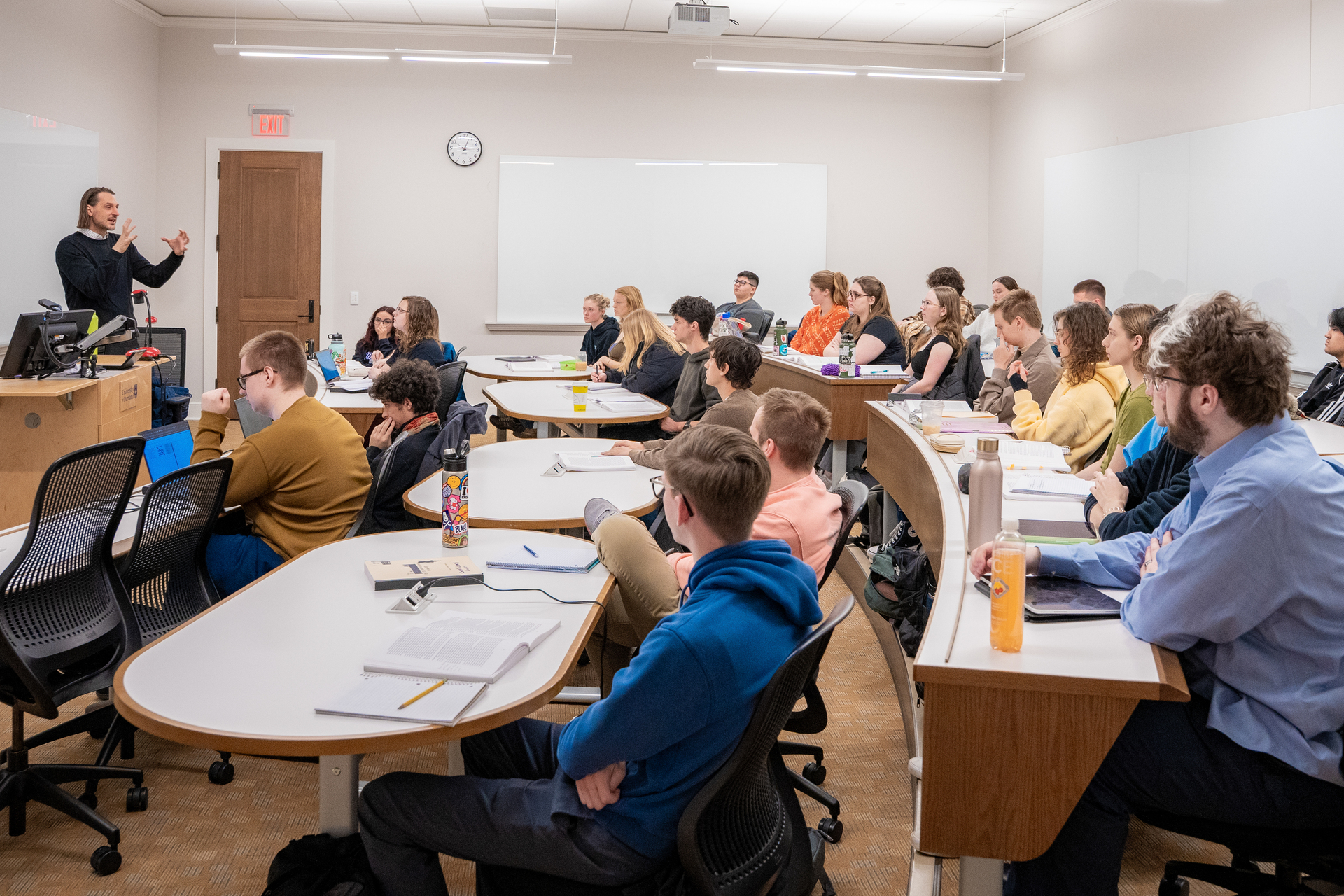 A group of University of Portland  students sits attentively , listening to a history lecture. Desks hold notebooks and personal items.