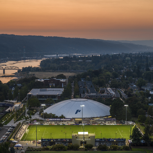 An aerial view of the Chiles Center and Merlo field at dusk. Lights illuminate a game on the soccer field with a rosy sunset glowing in the background.