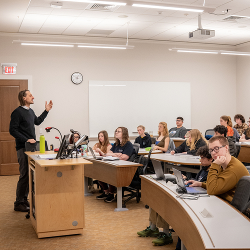 Students seated in curved tiered desks rows listening to a history lecture; some use laptops. A whiteboard is visible at the front of the classroom.