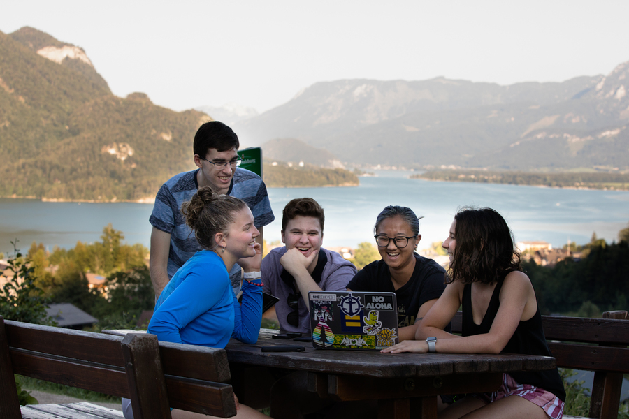 A group of University of Portland students sit at a picnic table while looking at a laptop computer with an alpine lake and mountains in the background.