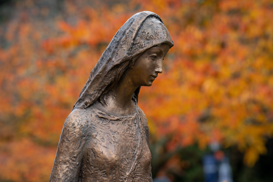 A close-up of the bronze statue of Mary in the grotto at the University of Portland against a blurred background of colorful fall leaves.