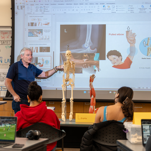 A UP professor stands in front of a digital projector screen while pointing to a model of a human skeleton during a kinesiology lecture.