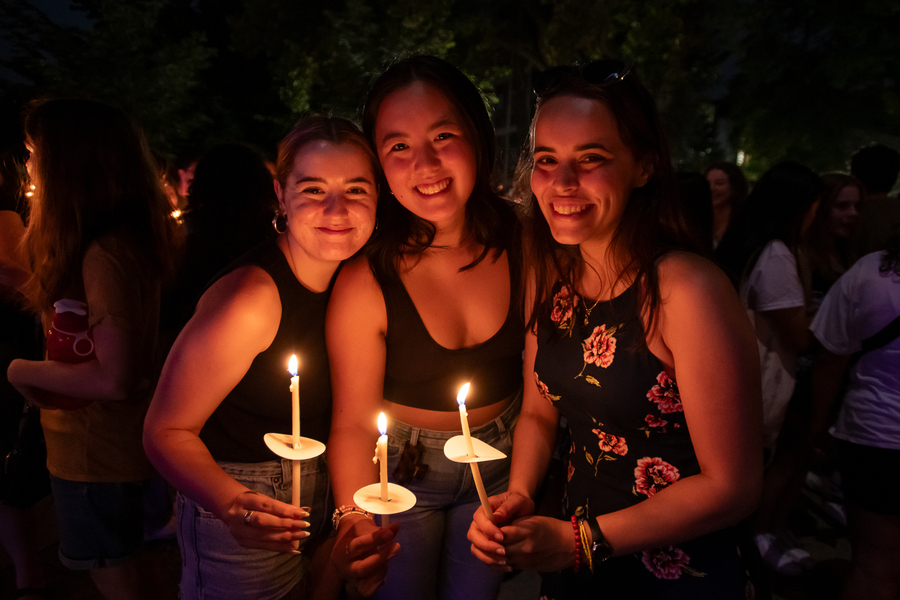 Three students smile and pose closely while holding lit candles at the bell tower candle ceremony at the University of Portland.