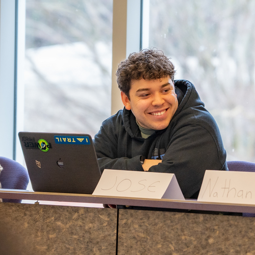 A UP undergraduate student smiles with casually crossed arms while seated behind a desk in front of their computer during a business class. Only the upper half of their body is visible.