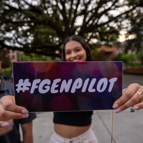 A first generation student at the University of Portland holds a small sign that says FGENPilot close to the camera as the focal point of the image.