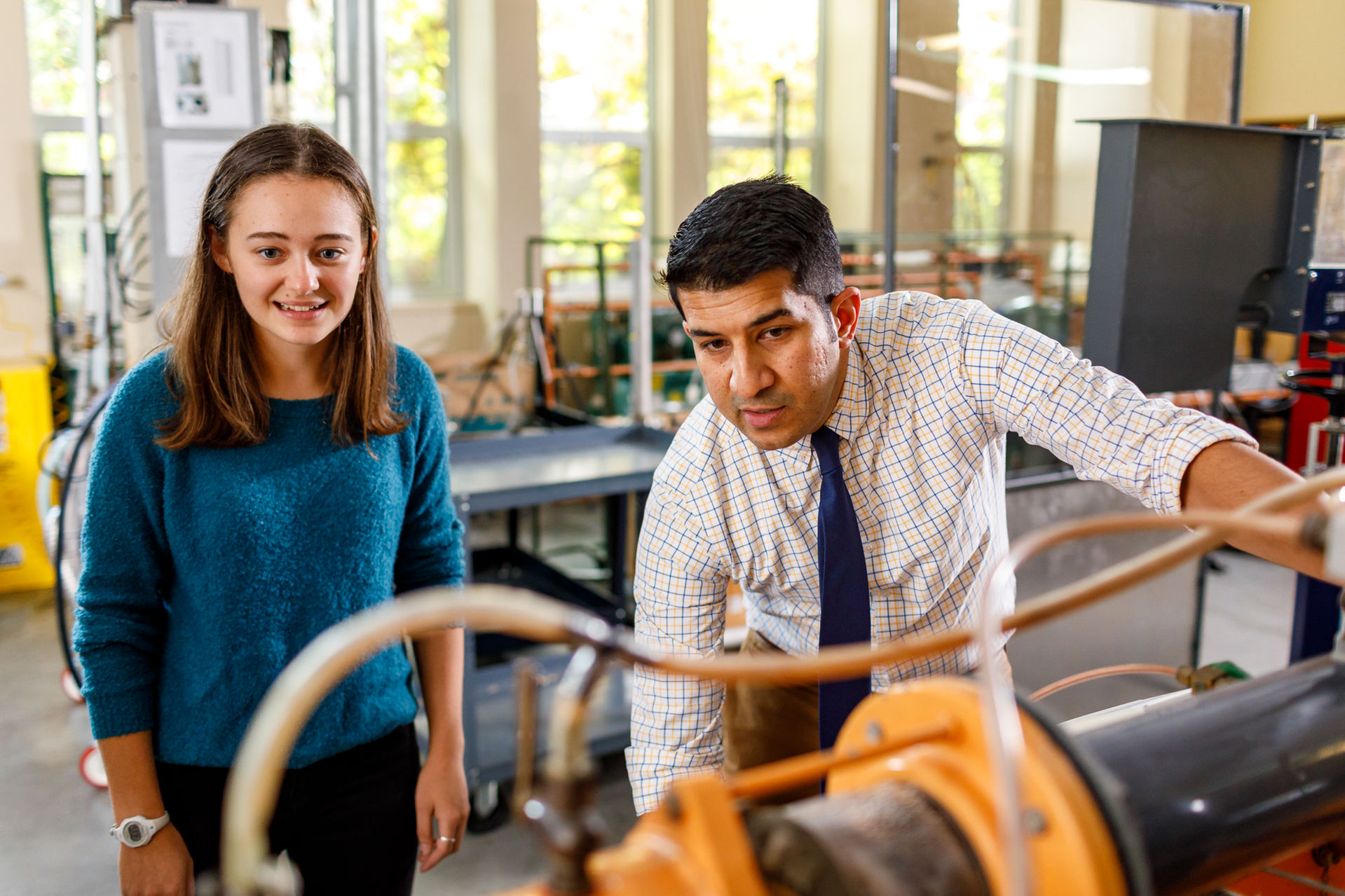 A UP student with long brown hair wearing a teal sweater stands next to a professor, who is adjusting a piece of lab equipment. The background is blurred and only their upper torsos are visible.