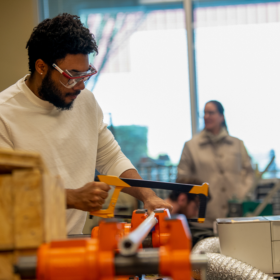 A University of Portland mechanical engineering wearing a pair of saftey glasses cuts a metal pipe with a hacksaw at a workbench in a workshop, with tools and equipment visible in the background.