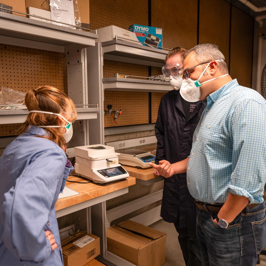 A mechanical engineering student in a blue lab coat, professor and another student in a black lab coat and mask examine laboratory equipment on a wooden shelf in a university of portland lab environment.