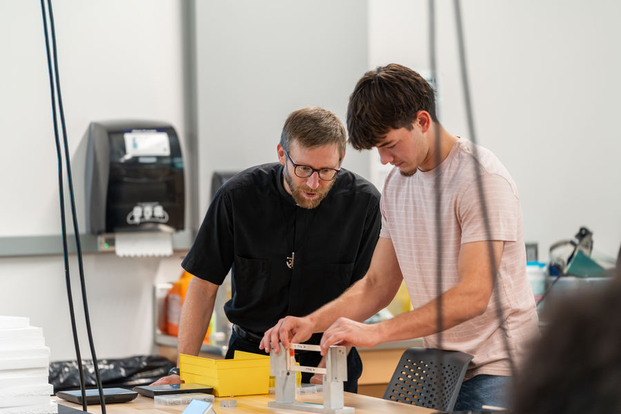  A UP mechanical Engineering student in a light pink, short-sleeved, striped shirt works on a project in the lab. His mentor, a Holy Cross brother is standing to his right instructing him. 