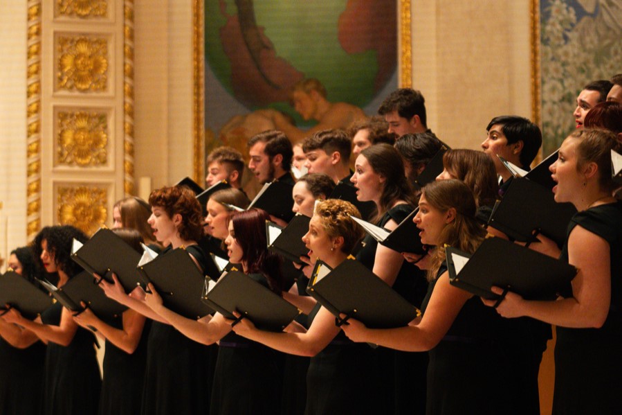 University of Portland students perform in a choir, holding sheet music, with a softly lit background and partially visible artwork behind them.
