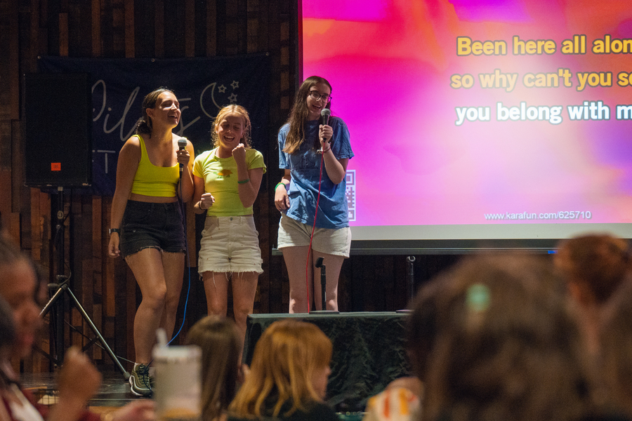 Three UP students stand on stage while singing karaoke in a night club against the back drop of a projector screen with lyrics at the Pilots After Dark event.