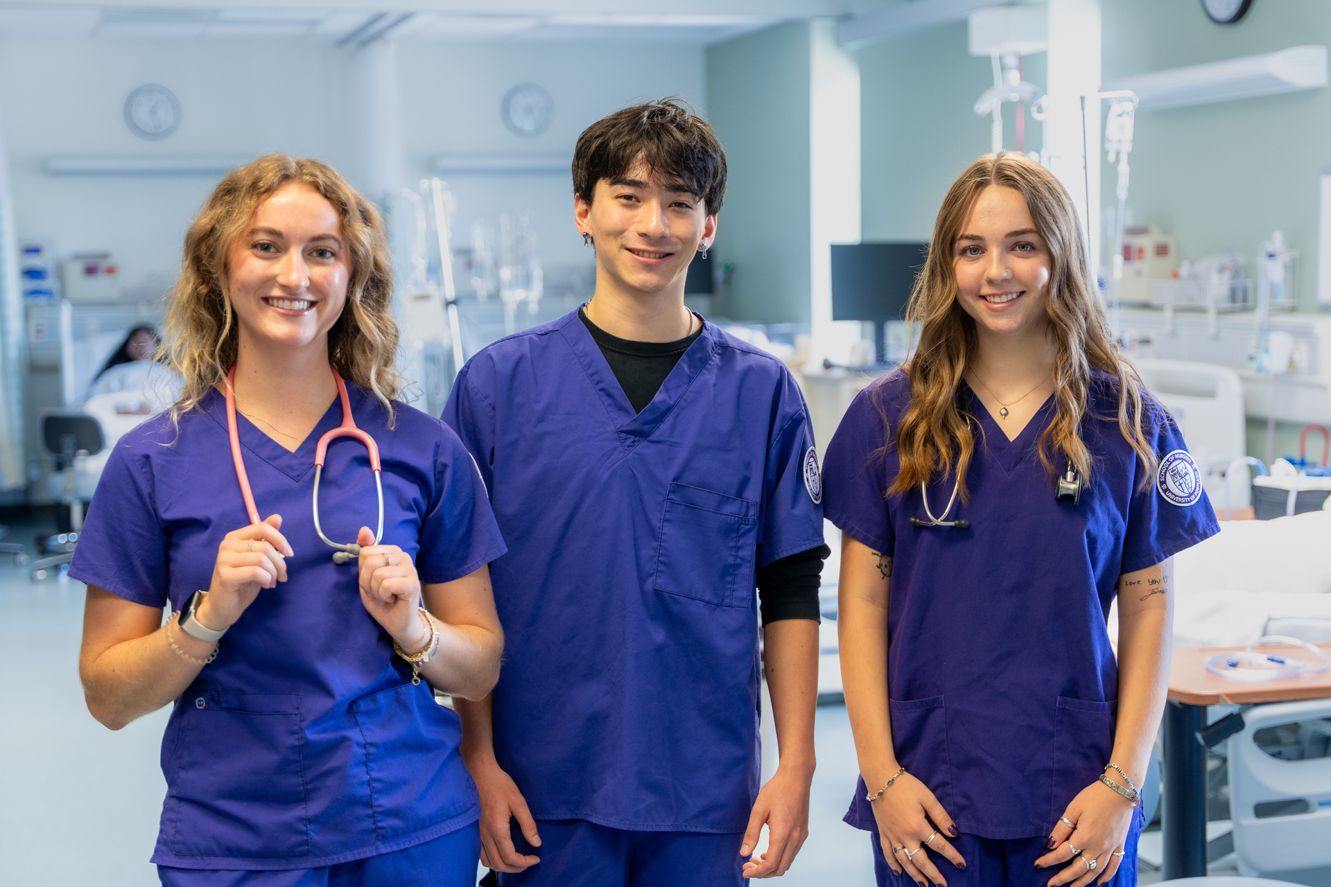 Three University of Portland nursing students in UP purple scrubs, two with stethoscopes, stand together with the clinic simulation lab in the background