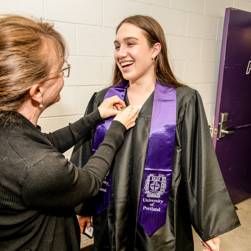 A University of Portland nursing student in graduation regalia smiles as their instructor affixes a special pin to their graduation robe.