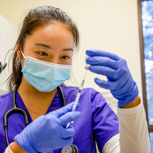 A close up image of a nurse wearing a mask and nitrile gloves, practicing injection techniques with a hypodermic needle and vial in the simulated health lab at the University of Portland.