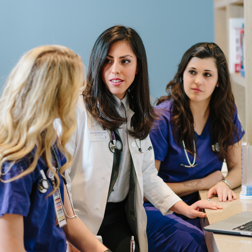 Two student nurses in purple scrubs flank their instructor, wearing a white medical coat, as she provides guidance in the simulated health lab at the University of Portland.
