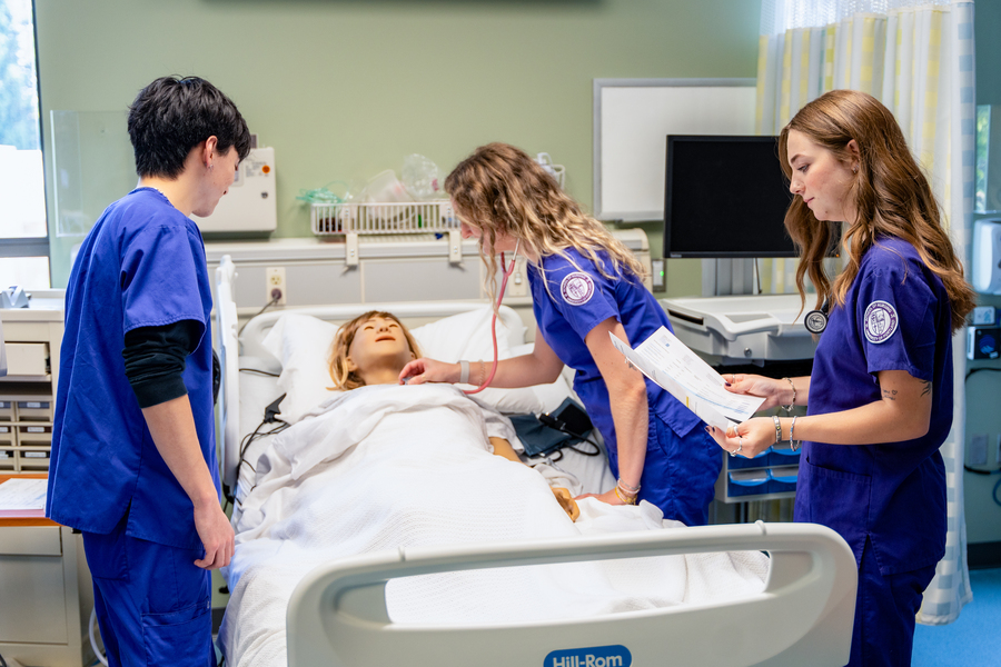 Three nursing students in purple scrubs attend a patient mannequin in the simulated health center at the University of Portland.