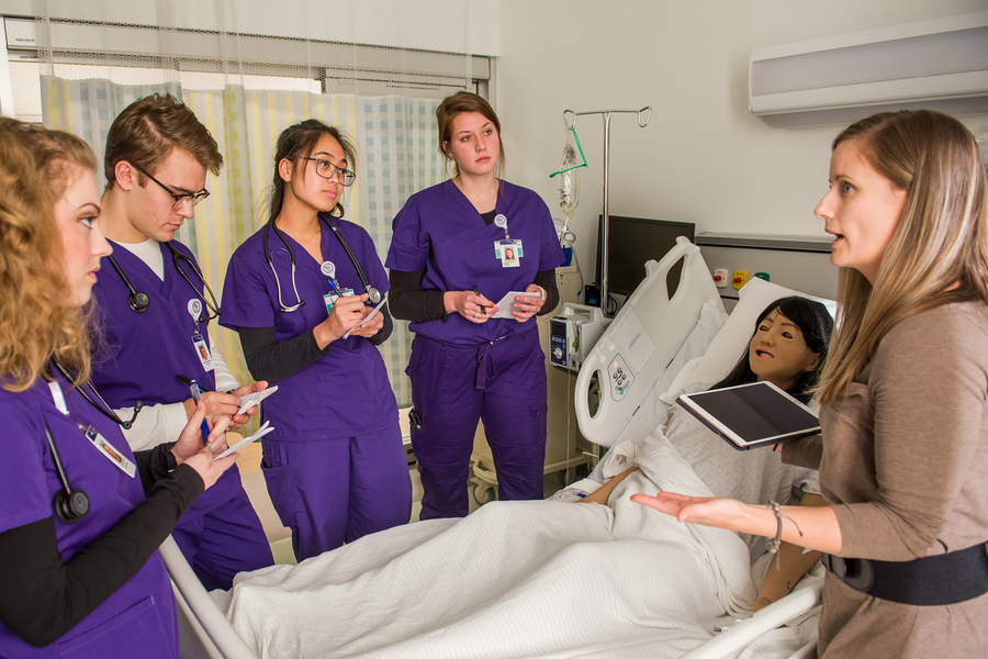 Nursing students in purple scrubs gather around a patient mannequin with their instructor as they practice bedside procedure in the simulated health lab at the University of Portland.