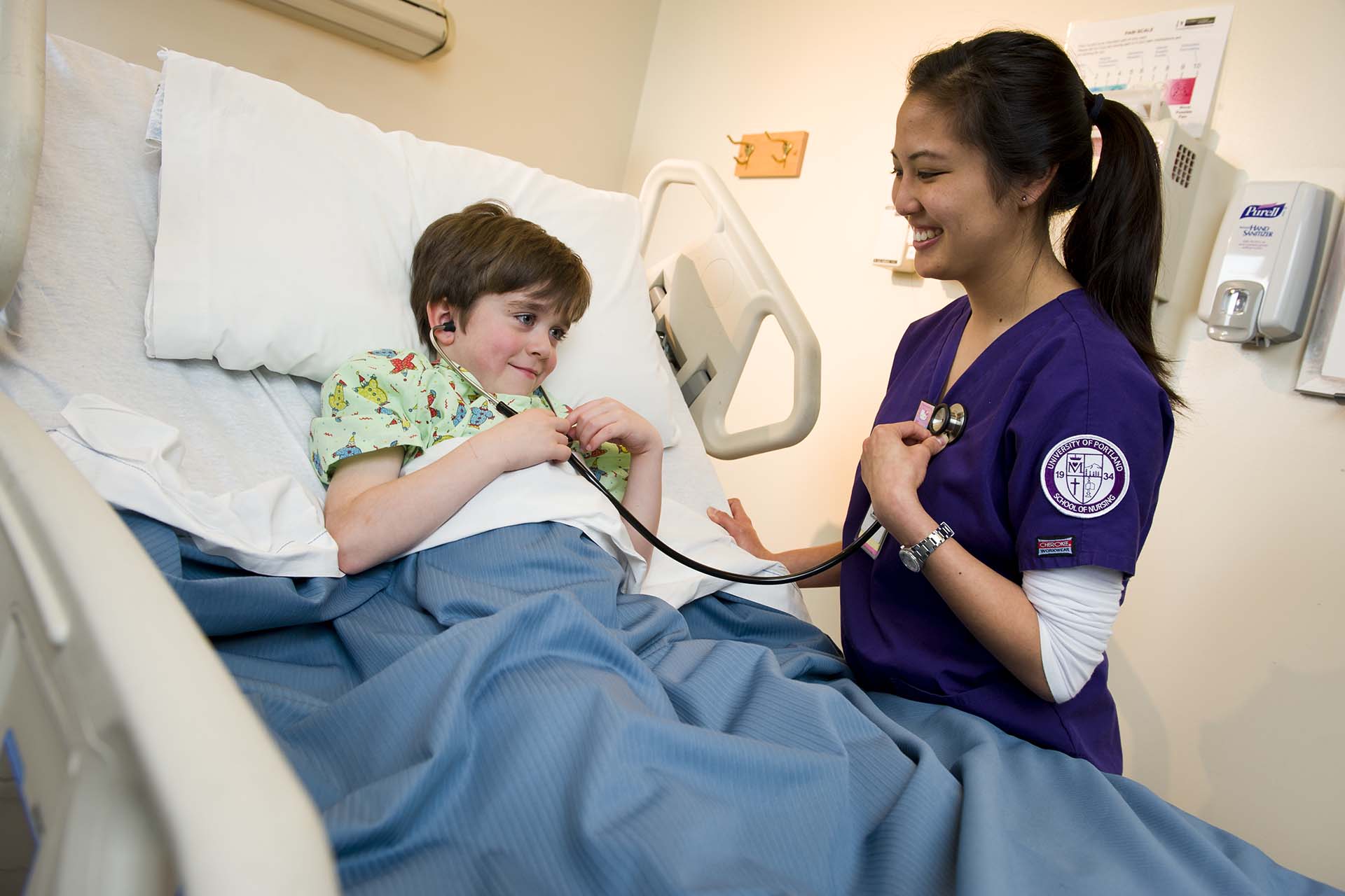 A University of Portland nursing student allows a young patient to listen to her heart with a stethoscope as the child lays in a hospital bed