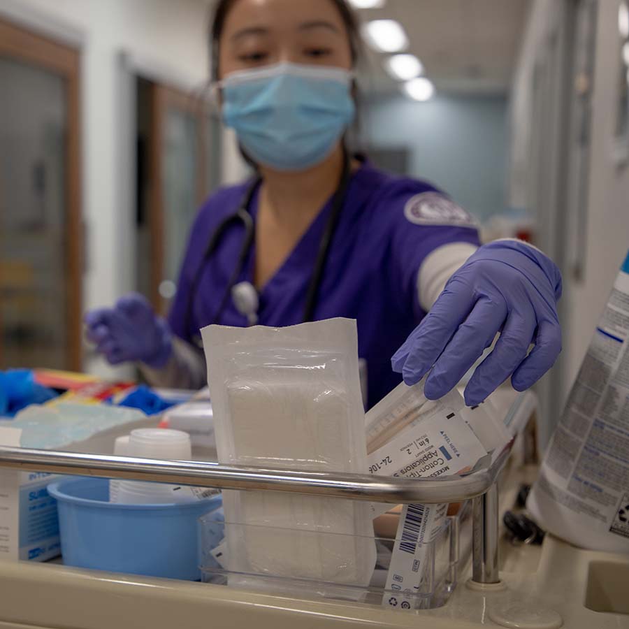 A University of Portland nursing student wearing a medical face mask selects medical supplies from the cart in the clinic simulation lab hall