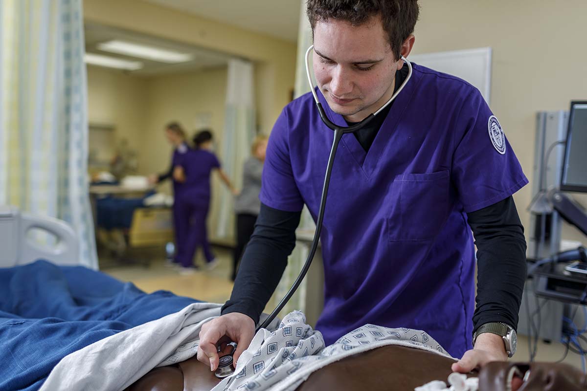 A University of Portland nursing student checks the vitals of a medical mannequin with a stethoscope in the clinic simulation lab while other students work in other simulation rooms in the background