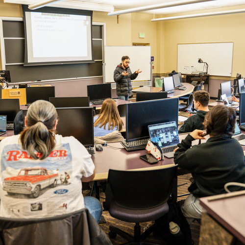 An instructor presenting to operations management students in a computer lab, with monitors, whiteboards, and office equipment visible in the background at the University of Portland.