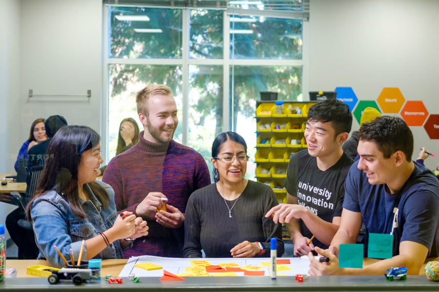A professor stands at the center of a group of students gathered in front of a table covered in colorful sticky notes as they brainstorm in the Innovation Lab at the University of Portland.