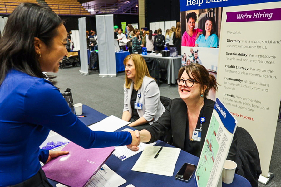 Two individuals shake hands across a table with a blue tablecloth, in the spacious indoor setting of the UP Career Fair.
