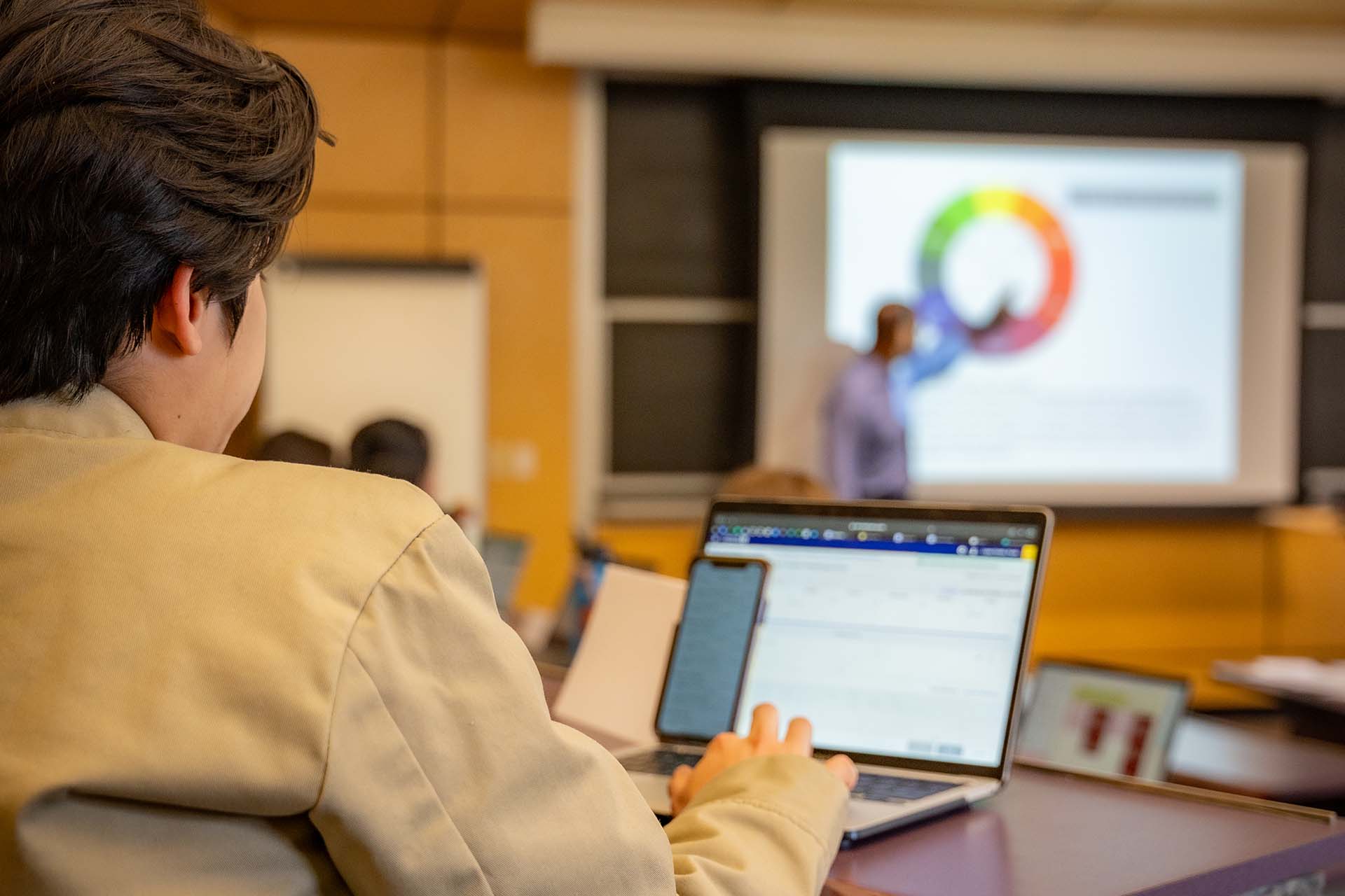 A University of Portland student takes notes on a laptop while professor points to a colorful pie chart on the projector screen in the background during a strategic management lecture