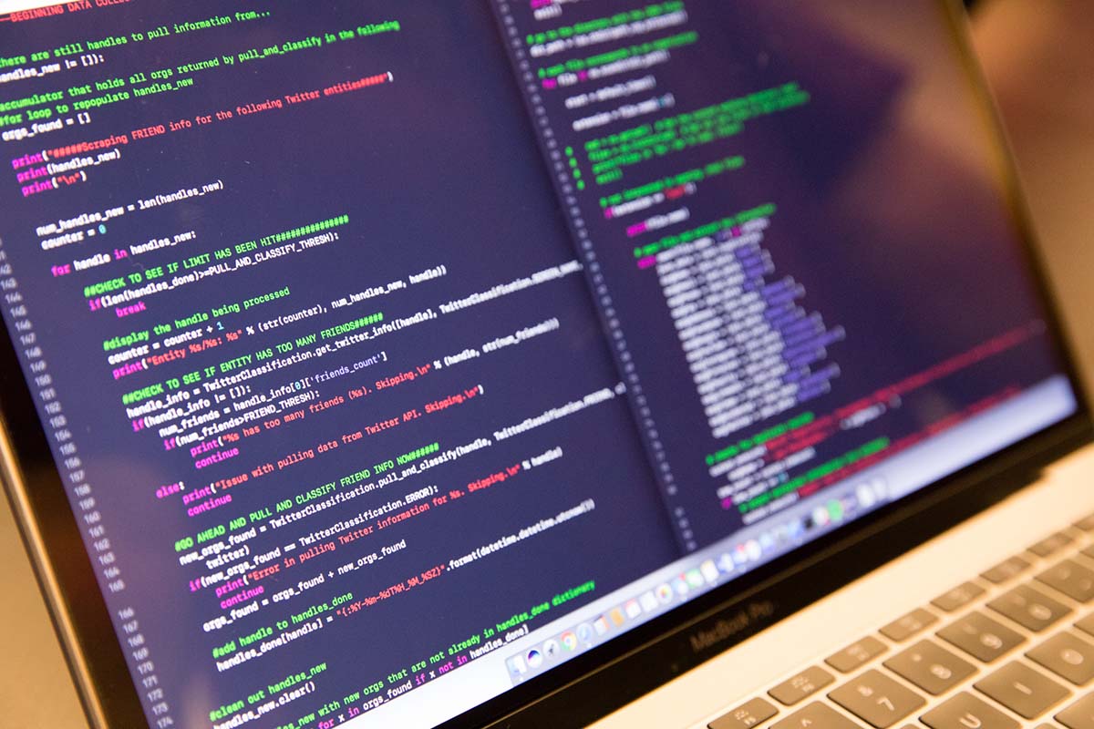 Close up of a silver MacBook with a black screen filled with multi-colored text in a coding program language
