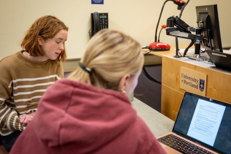 Two students, seated at a desk and viewed from behind, discuss a topic during an organizational communication class at the University of Portland.