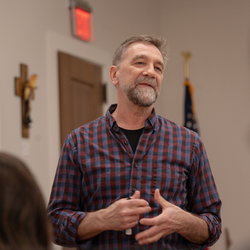 A professor, who is a middle-aged man with short gray hair and a beard, smiles slightly while teaching philosophy in the classroom at the University of Portland.