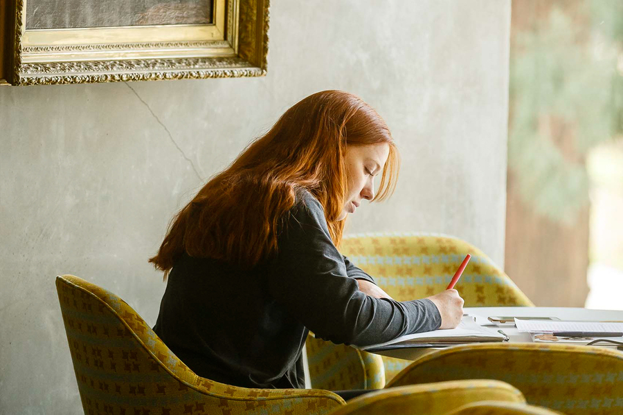 A student with long red hair sits and looks down thoughtfully as she studies, softly illuminated by natural side lighting in a quiet lounge at the University of Portland.
