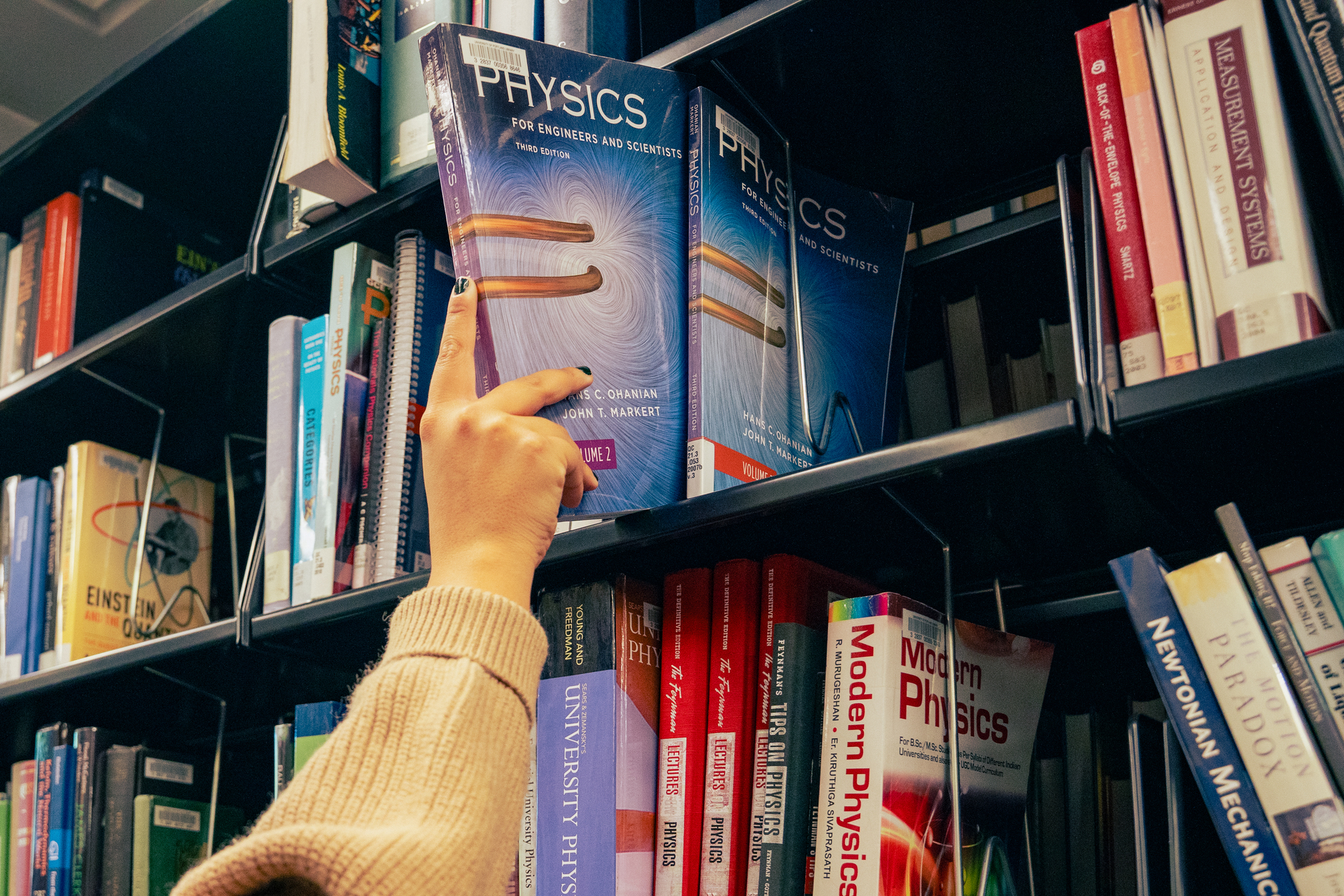 A  University of Portland student reaches up and grabs a Physics textbook off of a metal library shelf. The shelf houses a number of books with spines in red, yellow, and white covers, arranged in a geometric pattern.
