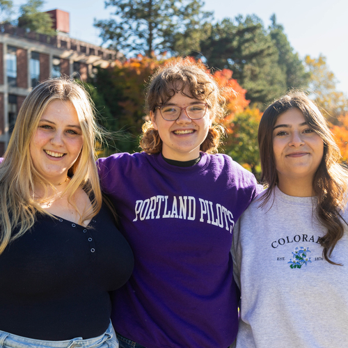 Three UP students smile outside on campus, standing in front of a university building and trees on a sunny day.