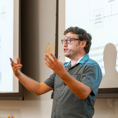 A University of Portland physics professor stands in front of a digital projection screen and gestures with his hands while speaking during a classroom lecture.