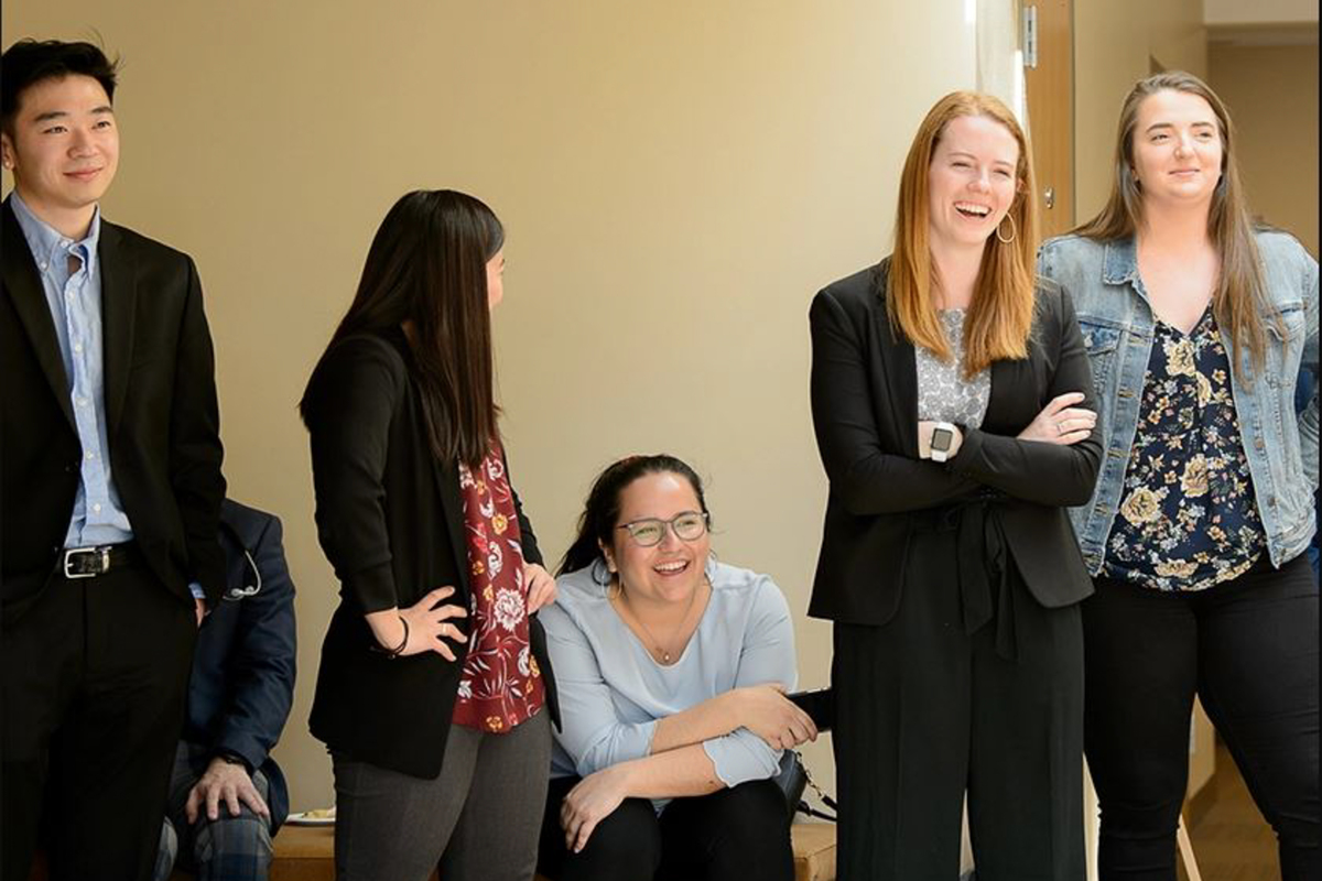 Five UP business students in professional attire are casually gathered in a hallway smiling expectantly before a presentation.
