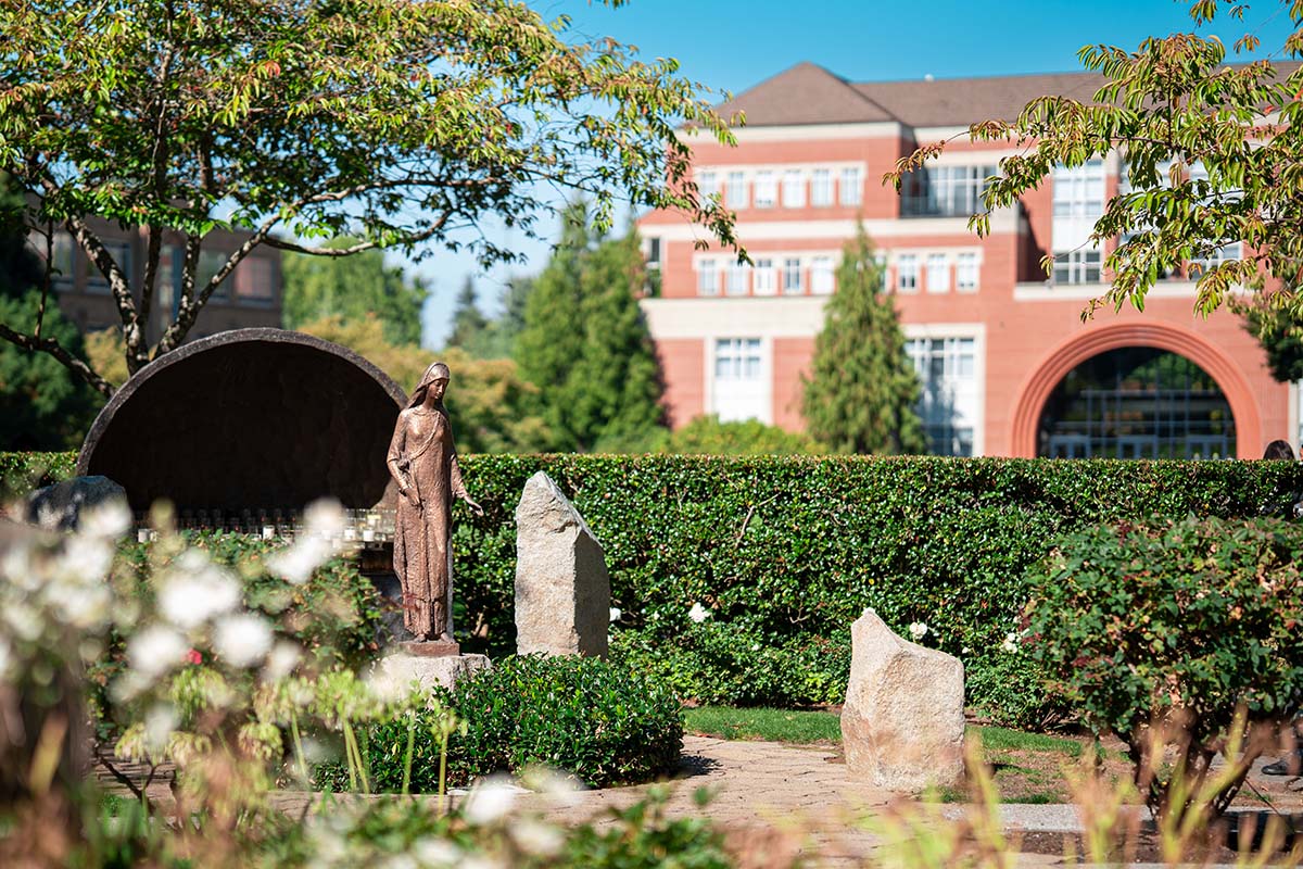 A picturesque photo of the Mary grotto on a sunny day with greenery and flowers and Franz Hall in the background