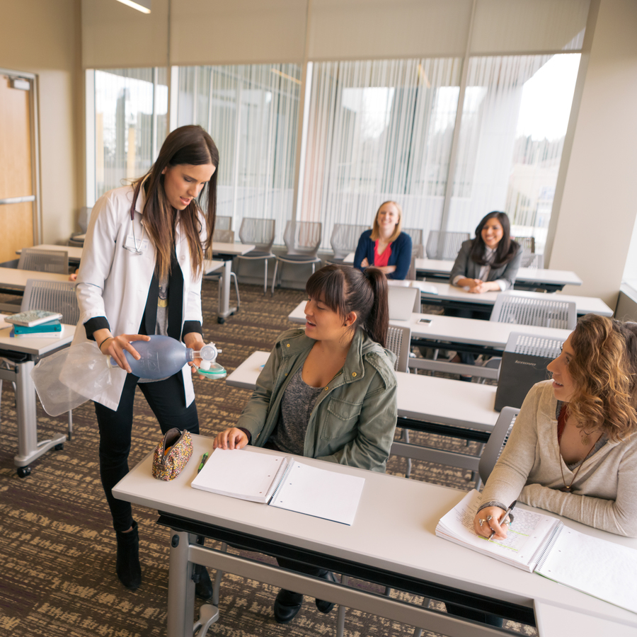 A University of Portland school of nursing professor demonstrating a manual resuscitator with a pediatric size valve mask to a student sitting at a table in one of the classrooms located in the simulation clinic