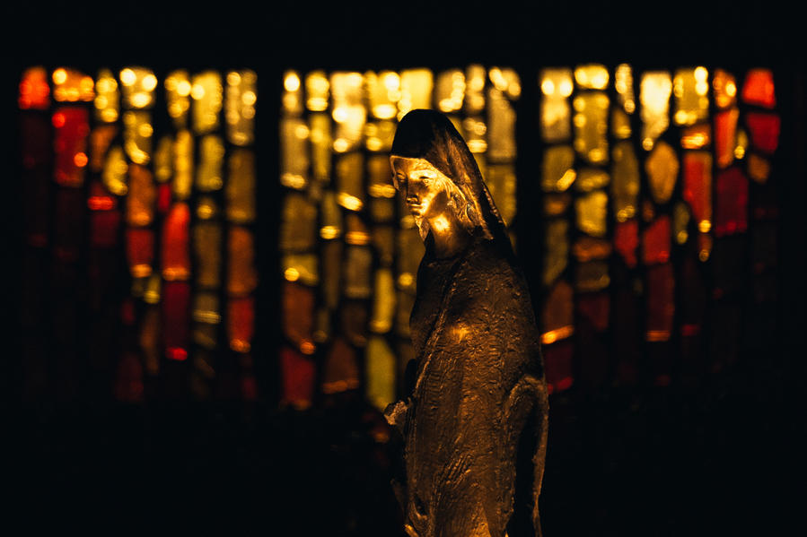 A night time image of the statue of Mary in the Grotto at the University of Portland. A stained glass window glows with warm colors in the background.