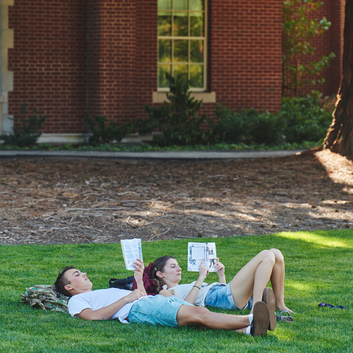Two students lie in the grass while reading books under the shade of a tree on the University of Portland campus on a sunny day.