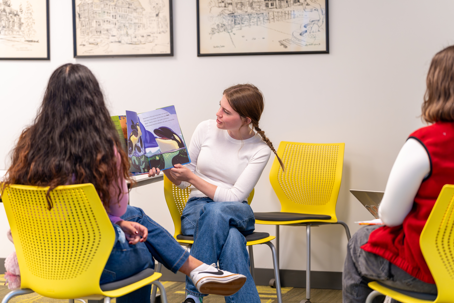 A woman in a white shirt and blue jeans presents a colorful picture book to another seated individual in an academic setting.