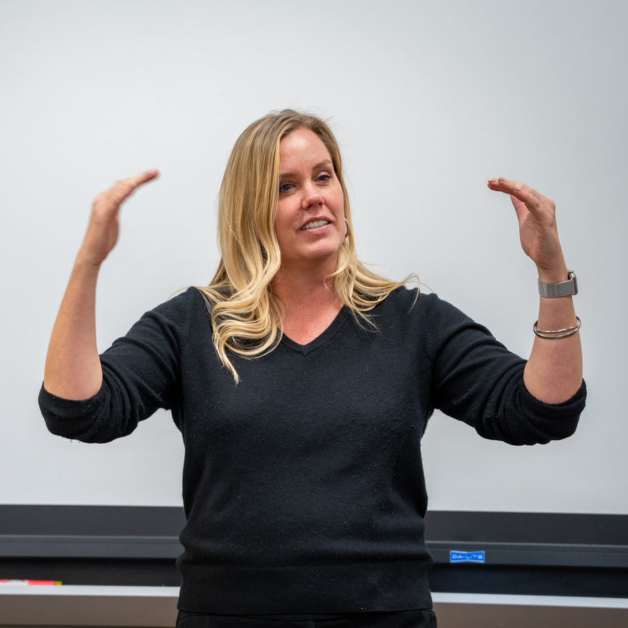 A University of Portland Communications professor stands in front of a white board with her arms raised as she explains communications and organizational hierarchy to her class