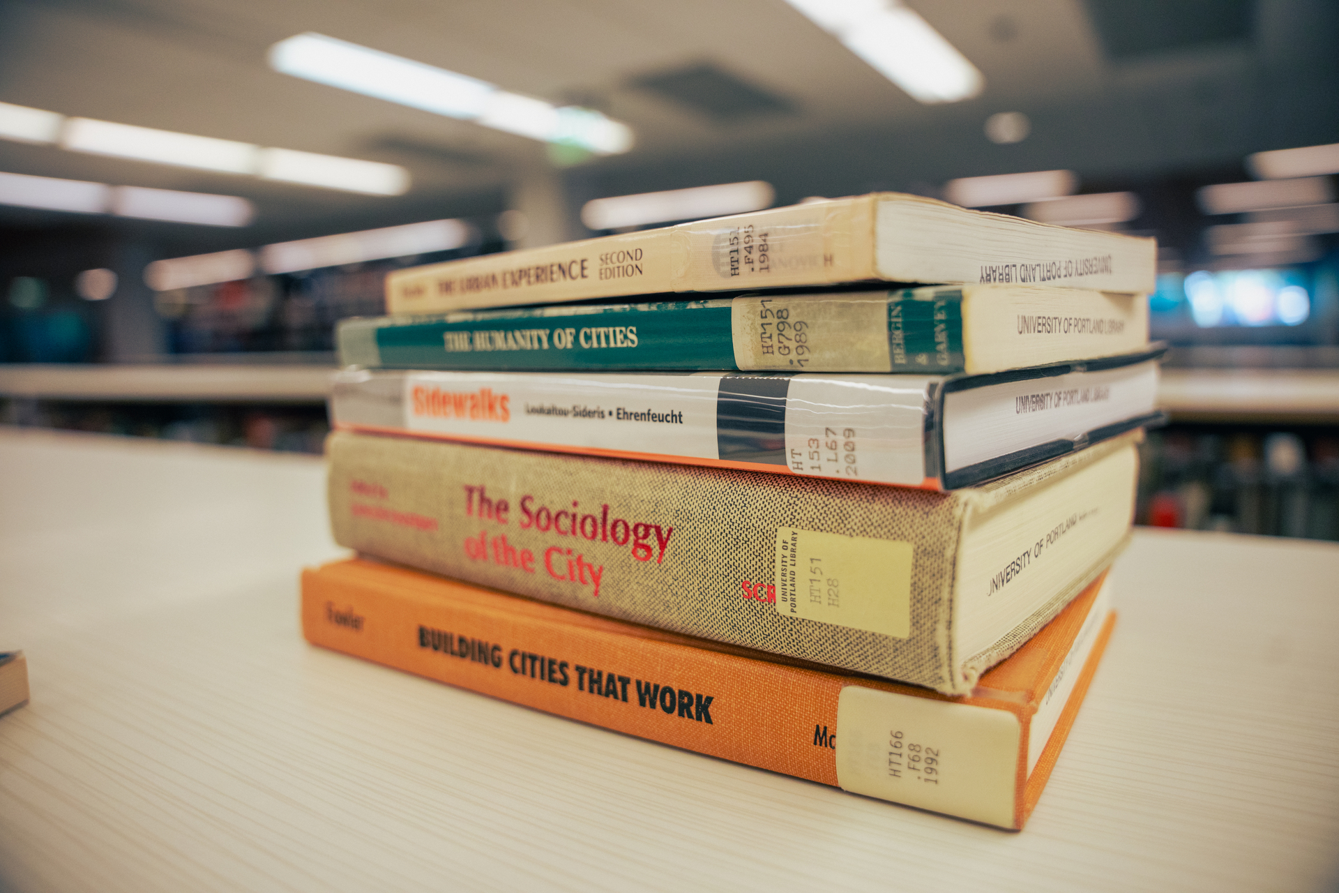 A close-up of stacked Sociology books, with the top one titled The Urban Experience Second Edition, and blurred library lights in the background.