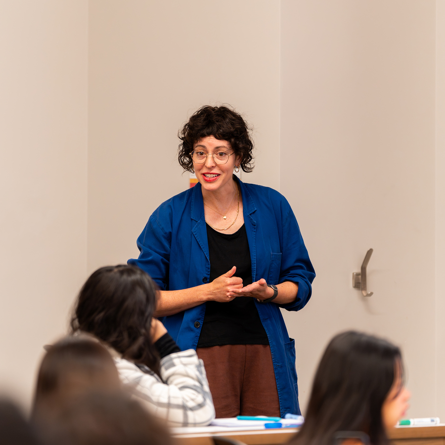 A UP professor with short, curly dark hair, glasses, and red lipstick smiles while looking slightly to the side while listening to student discussion in the sociology department's Understanding Society course. 