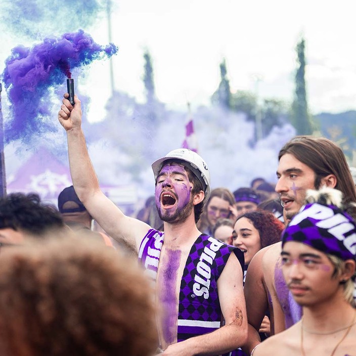Members of the Pilots drum squad covered in purple paint, wearing Portland scarves, and carrying purple smoke bombs march into the stadium with other fans