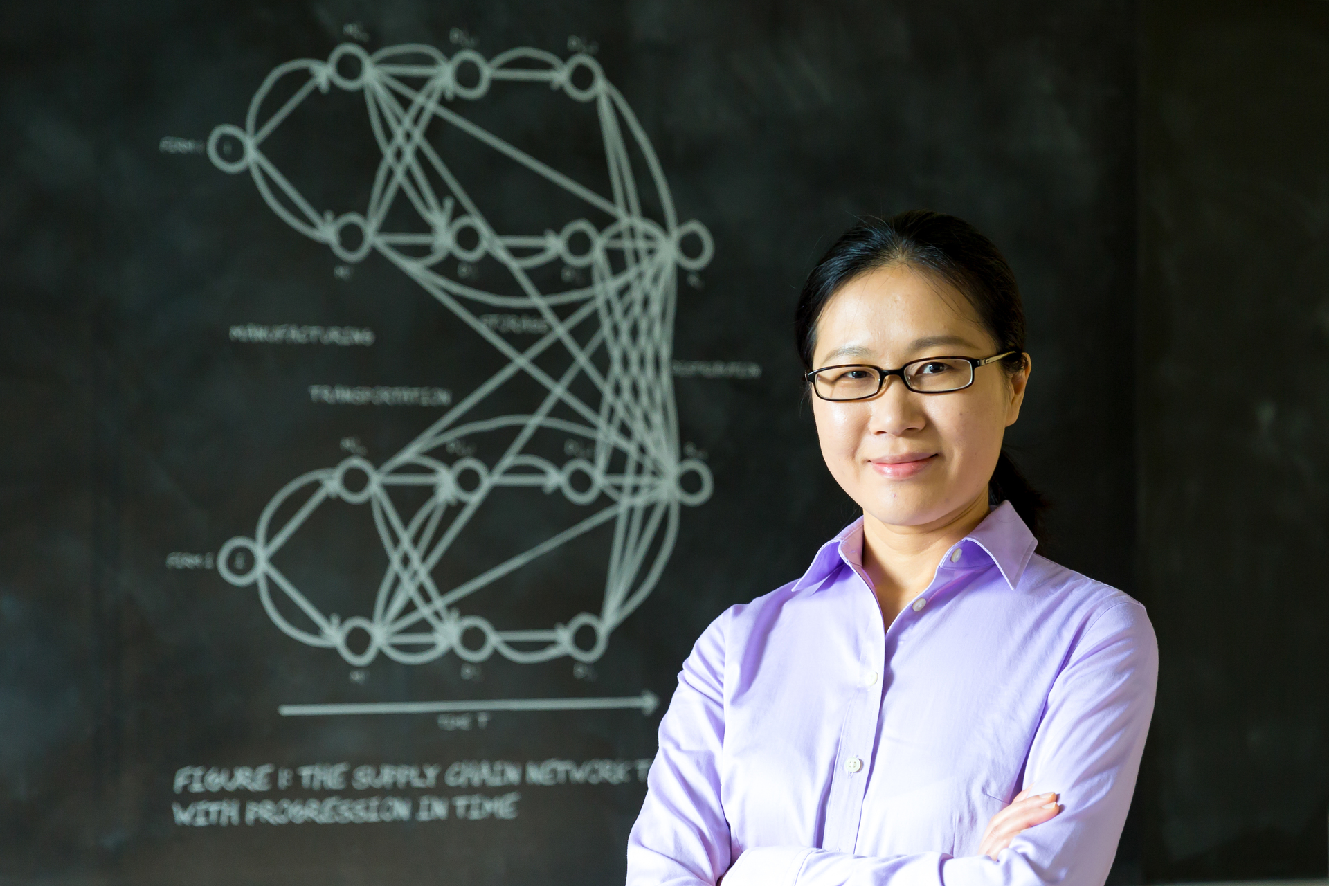 A smiling University of Portland supply chain analytics student stands with crossed arms in front of a process illustration on a chalkboard.